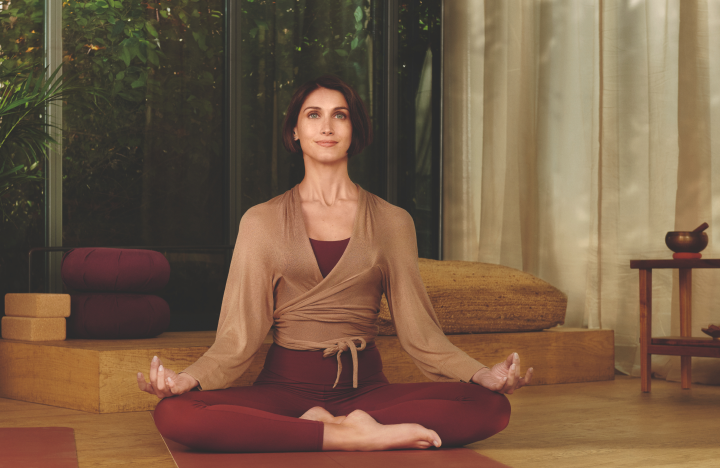  Egy rövid barna hajú nő ül törökülésben, békés meditációs pózban. Világosbarna, átkötős felsőt és bordó leggingset visel. A szobában nyugodt, természetes háttér veszi körül, növényekkel és lágy megvilágítással.