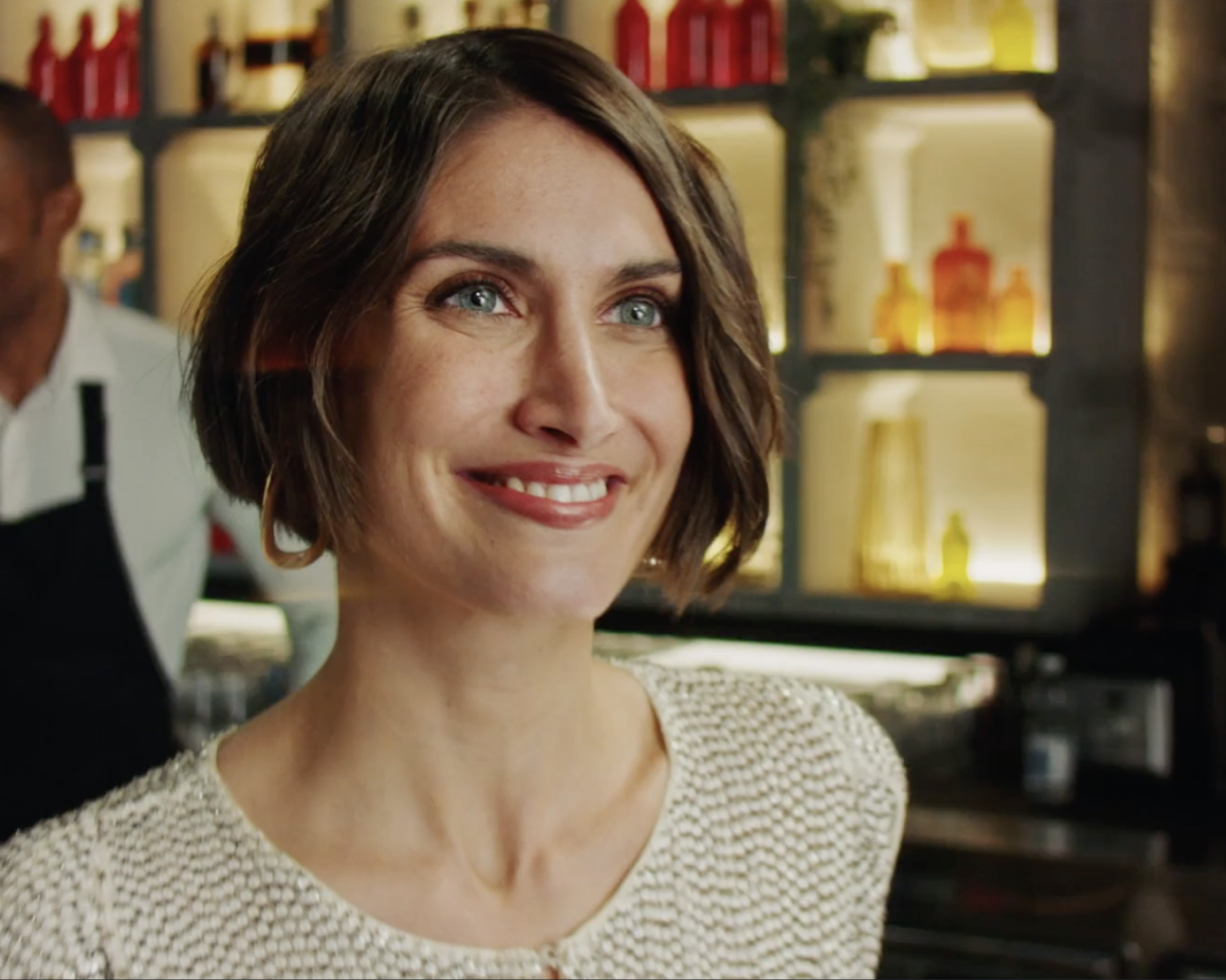 Smiling person with short brown hair, wearing a textured light sweater, standing in a warmly lit setting with shelves of bottles and decor in the background.