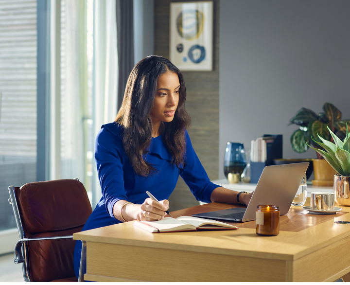 Woman in blue dress working in office looking at laptop and writing in notebook