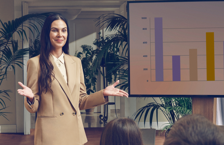 A woman with long dark hair, dressed in a beige blazer, stands in front of an audience giving a presentation with a bar chart displayed on a screen beside her. The setting includes large indoor plants and soft natural lighting.